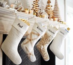 three stockings hanging from a mantel decorated with gold and white christmas decorations, along with candles