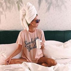 a woman sitting on top of a bed wearing a towel over her head and holding a cup