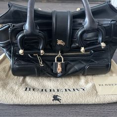 Authentic Burberry Purse Like-New Condition. Barely Ever Used. Black With Gold Hardware Features: Quilted Leather Body, Front Exterior Zip Pocket, Rolled Leather Handles, Front Strap With Padlock Closure, A Top Zip Closure, And An Interior Zip Pocket. Dimensions: Length: 14.5” Width: 6” Height: 8.5” Hand Drop: 12.00 Cm Shoulder Drop: 12.00 Cm Made In Italy Burberry Purse, Quilted Leather, Burberry Bag, Leather Handle, Zip Pockets, Burberry, Satchel, Bag Lady, Leather