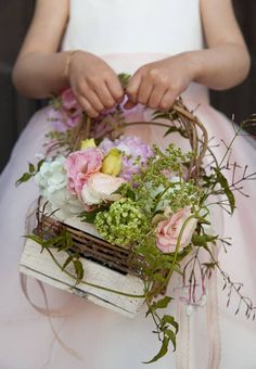 a woman in a white dress holding a basket with flowers and greenery on it