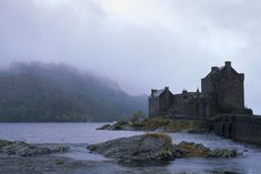 an old castle sitting on top of a mountain next to a body of water with fog in the air