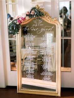 a wedding seating chart on a mirror in front of a door with flowers and greenery