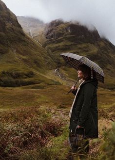 a woman holding an umbrella standing in the grass with mountains in the backgroud