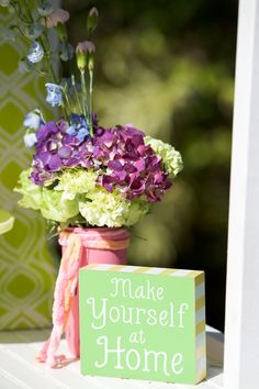 a vase filled with purple and green flowers next to a sign that says make yourself at home