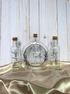 three glass bottles with writing on them are sitting on a gold satin tablecloth and there is a wooden wall in the background