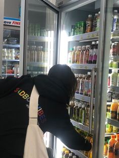 a person wearing a black hoodie looking into a refrigerator filled with drinks and juices