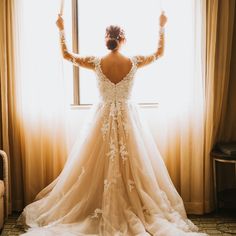 a woman standing in front of a window wearing a wedding dress and holding her arms up