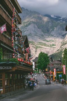 a town with mountains in the background and people walking on the sidewalk near buildings, cars and motorcycles