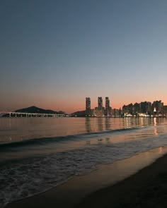 the city skyline is reflected in the water at sunset or dawn, as seen from the beach