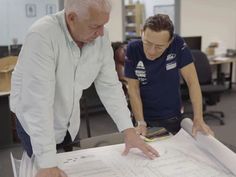 two men are looking at blueprints on a table in an open office space