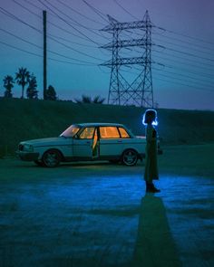 a person standing in front of a car at night with the lights on and an electric tower in the background