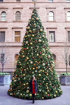 a woman standing next to a christmas tree in front of a tall building with lots of ornaments on it