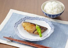 a white plate topped with fish next to a bowl of rice and chopsticks