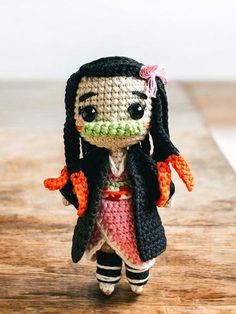 a small crocheted doll is standing on a wooden surface, wearing a black coat and pink dress