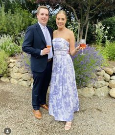 a man and woman standing next to each other in front of some bushes holding wine glasses