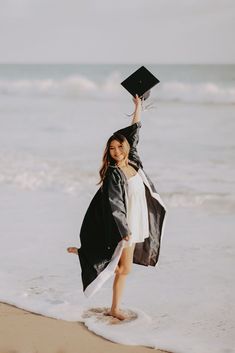 a woman in graduation gown holding up her cap and gown on the beach with waves crashing behind her