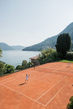 three people playing tennis on a clay court overlooking the water and hills in the distance