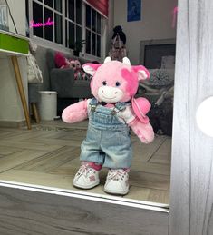 a pink teddy bear in overalls and white shoes standing up against a door frame