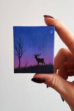 a hand holding up a small painting of a deer in the night sky with trees