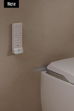 A close up of a beige wall. A white rectangular remote is on the wall. The corner of a toilet can be seen in the bottom right corner. A horizontal line of silver buttons is on the underside of the toilet lid. Smart Toilet, Smart Solutions, Toilets, New Generation