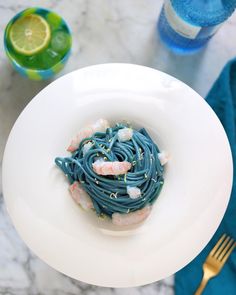 a white plate topped with blue pasta and shrimp next to a glass of lemonade