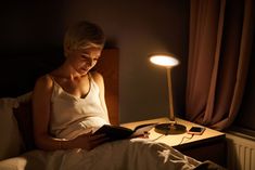 a woman sitting in bed reading a book under a light that is on top of a table