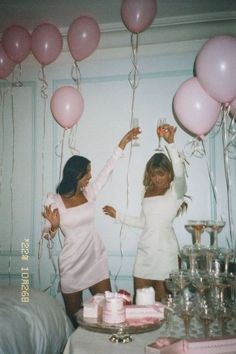 two women standing next to each other with balloons and cake on the table in front of them