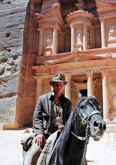 a man riding on the back of a black horse next to a tall stone building