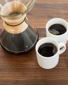 two cups of coffee sit next to an espresso maker on a wooden table