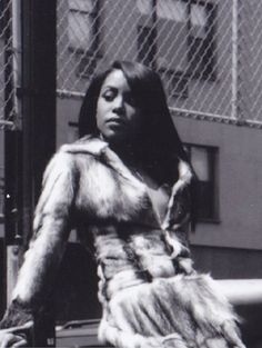 a black and white photo of a woman in fur coat standing next to a fence