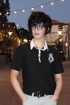 a young man with glasses standing in front of some string lights at night, wearing a black shirt and gray pants