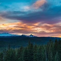 the sun is setting over some trees and mountain range in the distance with snow on top