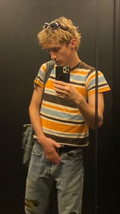 a young man standing in front of a mirror looking at his cell phone while wearing ripped jeans