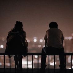 two people are sitting on a railing looking at the city lights in the distance,