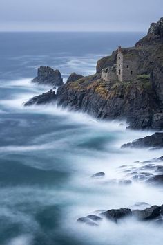 an old building on the edge of a cliff by the ocean with waves crashing around it