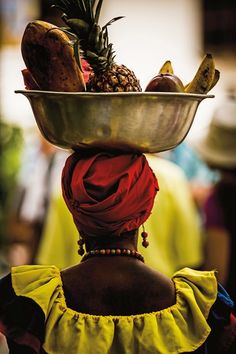 a woman with a large metal bowl on her head filled with fruits and veggies