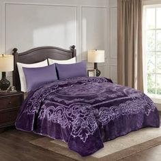 a bed with purple bedspread and pillows in a room next to a window