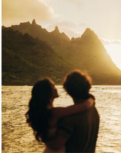 a man and woman standing in front of the ocean with mountains in the background at sunset