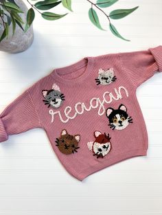 a pink sweater that says reason with bears and cats on the front, sitting next to a potted plant