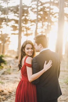 a man in a suit and woman in a red dress embracing each other while the sun shines through the trees behind them