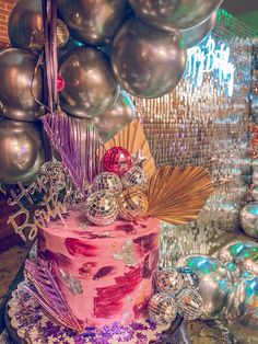 a pink cake with silver balloons and decorations on it's top, sitting in front of some disco balls