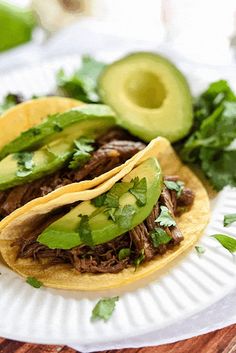 two tacos with meat, avocado and cilantro on a paper plate