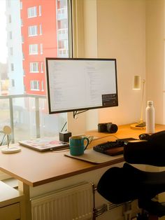 A minimalist home office setup featuring a large desk with a monitor, keyboard, and personal items. The workspace is bathed in natural light from a large window overlooking urban apartment buildings, creating a bright and inviting atmosphere. Minimal Aesthetic Home, Small Modern Apartment, Minimal Desk Setup, Male Living Space, Aesthetic Home Office, Modern Apartment Interior, Dream Desk, Computer Desk Setup