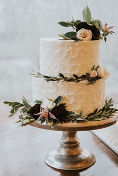 a wedding cake with white frosting and fresh greenery on top is sitting on a gold pedestal