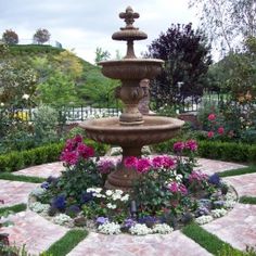 a water fountain surrounded by flowers and greenery in the middle of a stone walkway