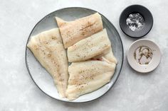 three pieces of fish on a plate with seasoning and salt next to one bowl