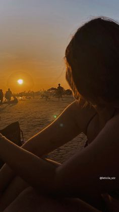 a woman sitting on top of a beach next to the ocean at sun set with people in the background