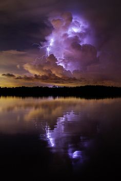 the sky is filled with purple clouds as lightning strikes in the distance over water and trees