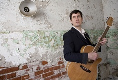 a man holding an acoustic guitar in front of a wall with peeling paint on it