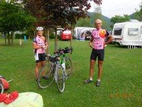 two people standing in the grass with bikes and campers behind them at a campsite